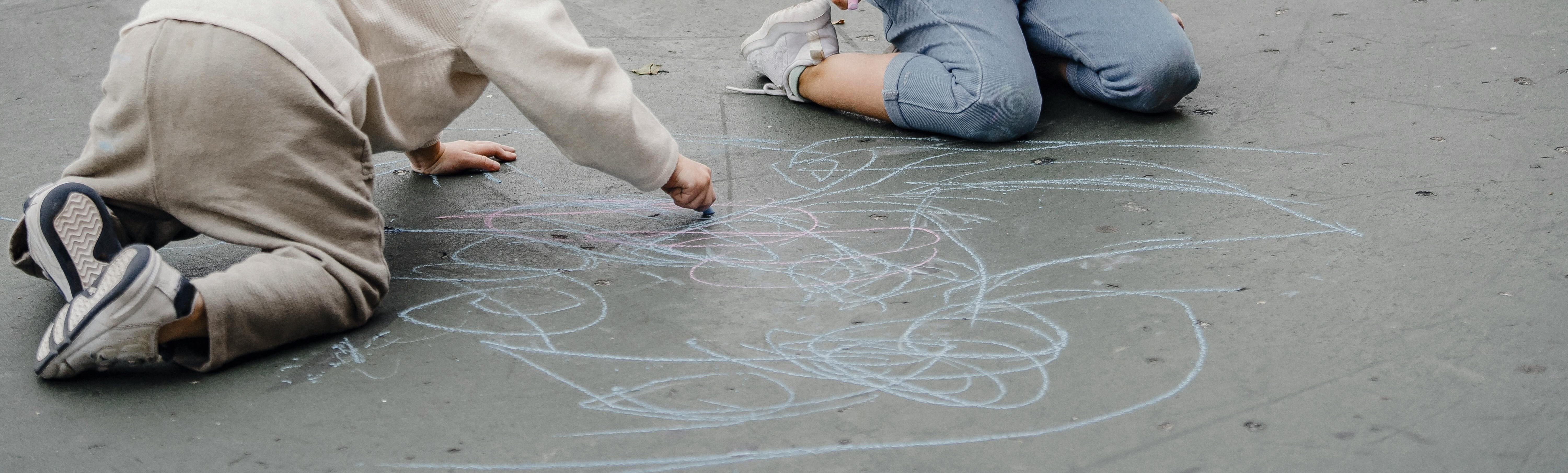 Two children are sitting on the ground drawing with colorful chalk. One child is focused on drawing on the pavement, while the other watches attentively. Various chalk drawings are visible around them.
