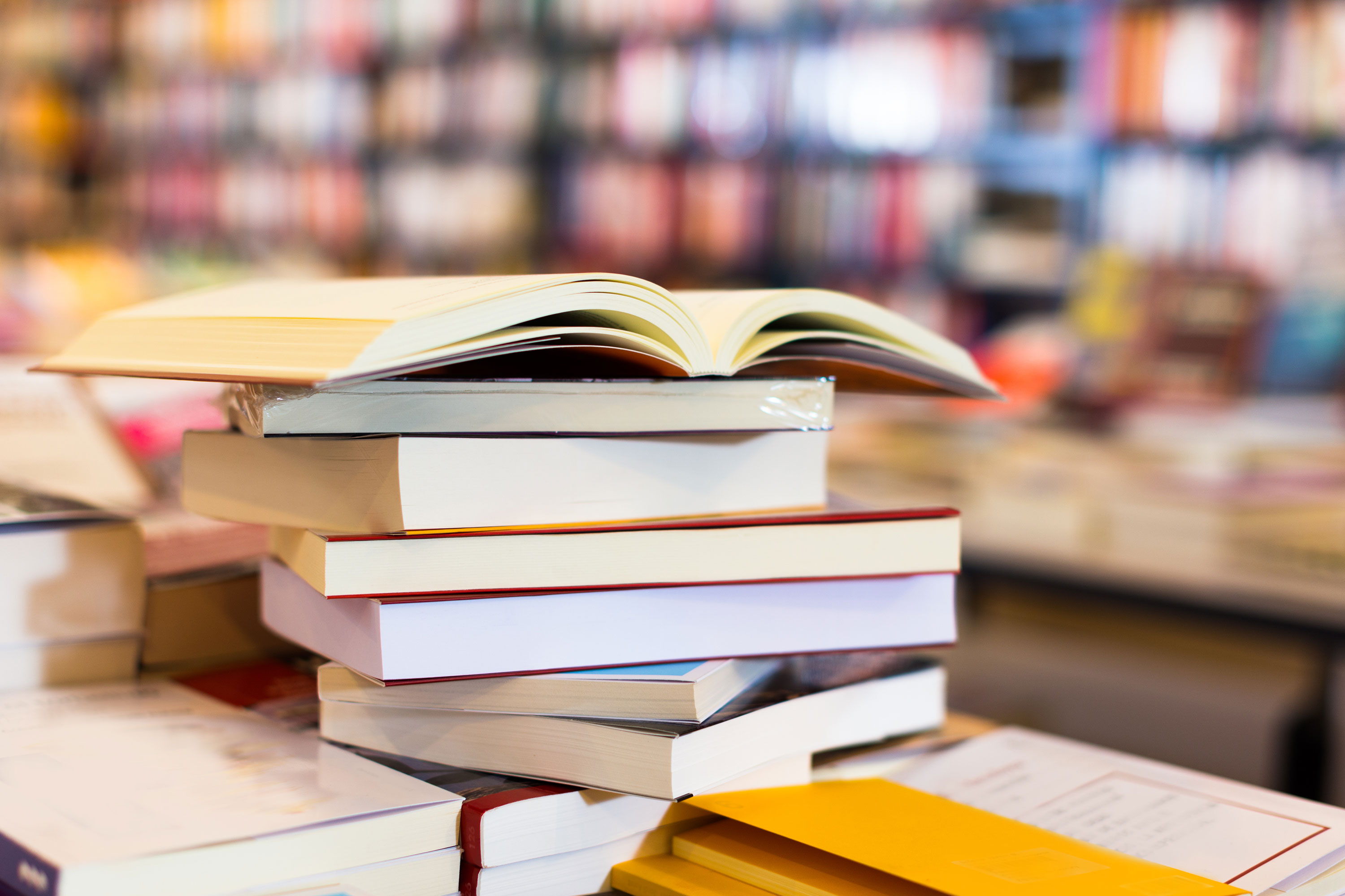 Ein Stapel Bücher liegt auf einem Tisch, mit einem aufgeschlagenen Buch obenauf. Im Hintergrund sind Bücherregale mit unscharfen, farbenfrohen Buchrücken zu sehen, was eine gemütliche Bibliotheks- oder Buchladenatmosphäre vermittelt.